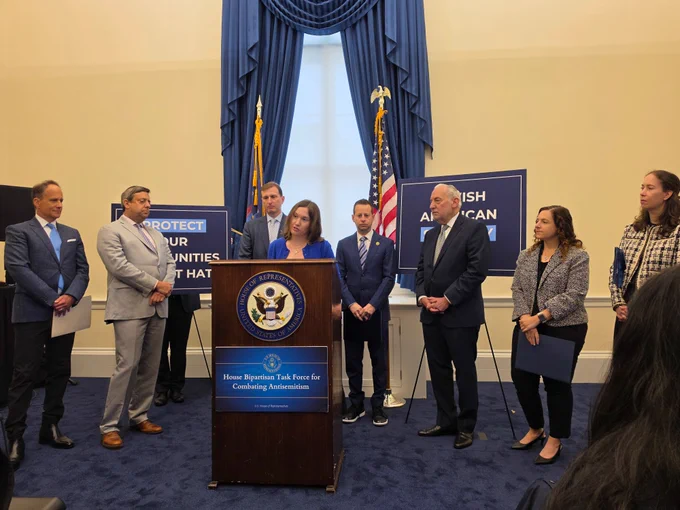 Amy Spitalnick, CEO of JCPA, speaks at a podium during a press conference on Capitol Hill alongside lawmakers and Jewish community leaders. Signs in the background reference the Jewish American Security Act and efforts to protect communities and combat antisemitism.