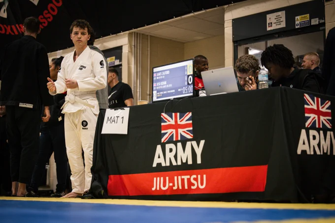 A person in a martial arts gi stands near a table marked "Mat 1" at an Army Jiu-Jitsu event. Two individuals sit behind the table with a computer and water bottles. A digital scoreboard is visible in the background.