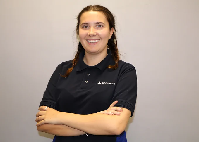 A photo of a female STS Defence apprentice stood against a pale wall. She is facing the camera smiling with her arms folded.