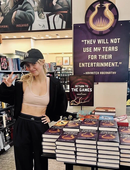 McKenna Grace poses in a Barnes & Noble bookstore wearing a black baseball cap, black jacket, and light pink top. She stands beside a Sunrise on the Reaping display featuring stacks of books and a large purple sign with a quote from Haymitch Abernathy.