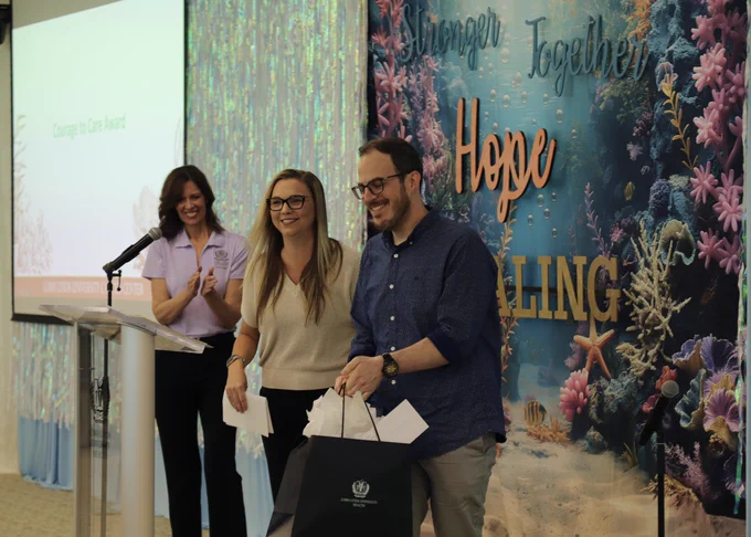 Two woman and one male are at an event. The backdrop features an ocean theme with coral and the phrases "Stronger Together" and "Hope and Healing." Two individuals stand together holding envelopes, one of them holding a gift bag. Another person claps while standing at a podium.