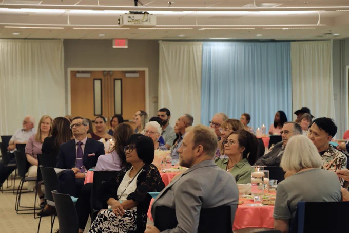 A group of people sit attentively in a decorated room with coral tablecloths and rows of chairs. The environment is a formal gathering and presentation, with a screen at the front.