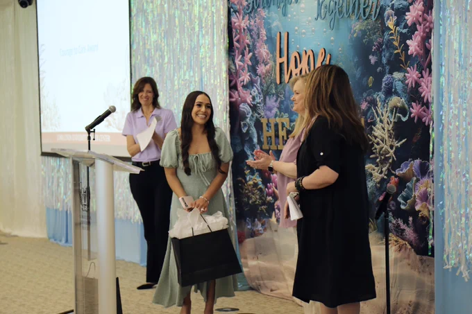 Four woman stand in a decorated room. One person at a podium smiles, while another person nearby holds a black gift bag smiling. Behind them, there is a colorful backdrop with the text "Stronger Together" and "Hope and Healing." and ocean-themed decorations.