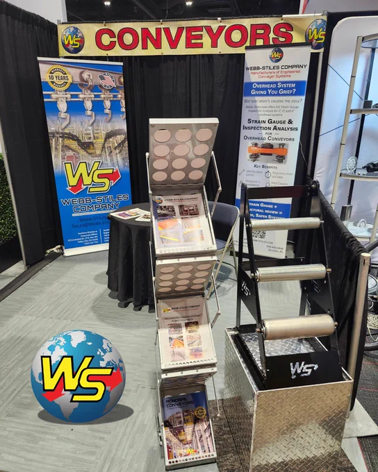 Webb-Stiles Company booth display at a previous FABTECH appearance, featuring the branded "Conveyors" banner, WS roll-up banner, a literature rack stocked with product brochures, and a roller conveyor demo unit on a diamond plate base. The Webb-Stiles logo globe graphic is featured in the foreground.