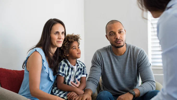 A group of three people, including a child, are seated on a couch, attentively listening to a person opposite them. The room has a neutral setting with a window visible in the background.