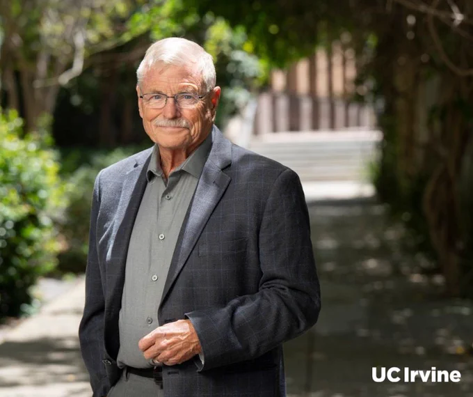 UC Irvine neuroscientist Oswald Steward stands outdoors on a tree-lined path. He has short gray hair and glasses and wears a gray shirt and a dark checkered blazer. The logo "UC Irvine" is visible in the bottom right corner.