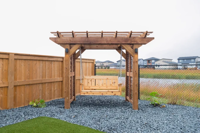 Wood pergola with a hanging swing bench installed in a landscaped backyard with decorative rock, fencing, and open green space in the background.