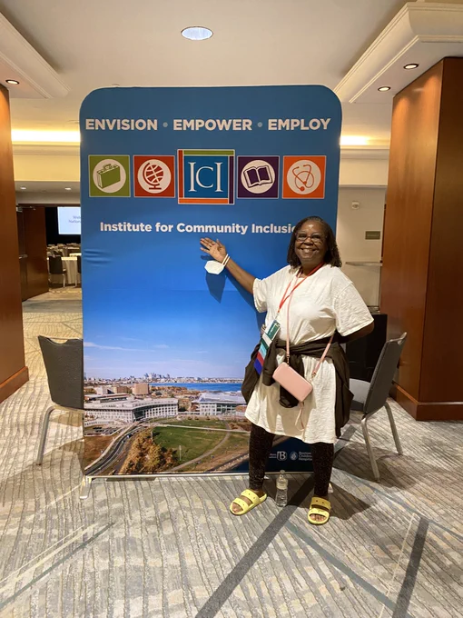 A Black woman in glasses smiles proudly while pointing at the ICI banner.