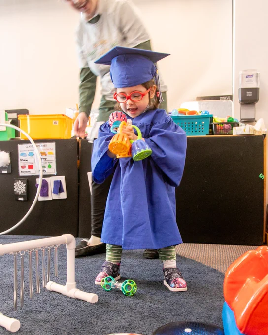 Isabella wearing a blue cap and gown, and bright red glasses, playing with a toy in a classroom
