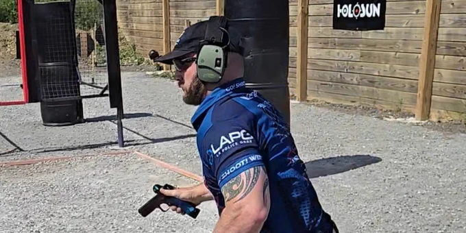Shooter in blue shirt and black shorts running on gravel at an outdoor shooting range with ear protection and handgun in hand.