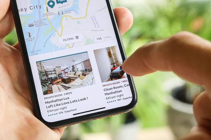 A person looking at listings for travel. Copy on the phone reads, "Manhattan Lux Loft. Like. Love. Lots. Look !"