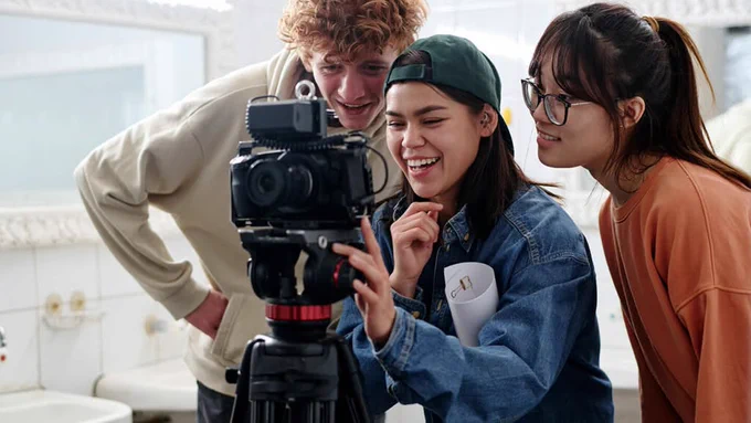 Three young adults closely examining a camera on a tripod in a bright room, discussing the shot setup.