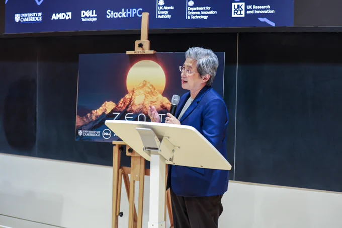 Dr Lisa Su in a blue blazer speaks at a podium in a lecture hall. Behind her is a poster with a mountain and the word "Zenith". The screen, also behind her, displays a group of logos.