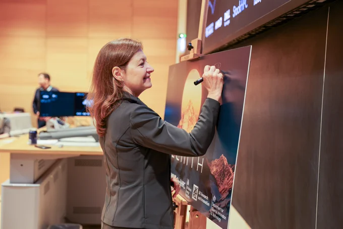 Professor Deborah Prentice is writing her signature on a large "Zenith" poster situated on an easel, with a classroom or lecture hall setting in the background.