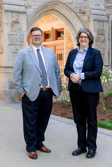 Two professionally dressed individuals standing in front of a building with stone architecture and an arched entrance. There are plants visible in the background.