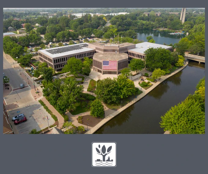 The City of Naperville logo and an aerial view of the City of Naperville Municipal Center facing the Riverwalk with an American flag on the building facing the DuPage River.