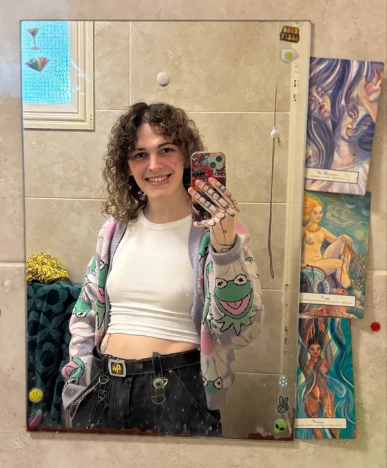 A picture of me taking a bathroom mirror selfie. Three small posters are propped up on its left side. I am wearing a white vest, black jeans and a purple fleece with kermit the frog’s face on it.