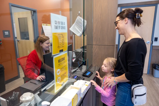 A woman and a young girl stand at a reception desk with a protective glass shield. The woman, dressed in black, has her hair tied up and a small crossbody bag. The girl, wearing a pink top, looks up at the receptionist, who is typing on a computer. The desk displays signs, including one about co-pay due at the time of service. The reception area has a modern design with a door marked "Break Room" visible in the background.