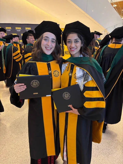 Two graduates in black and yellow academic regalia and hats, holding diplomas labeled "Iowa," smile for a photo. They stand in a crowd of similarly dressed graduates, in a bright indoor setting featuring university banners in the background.