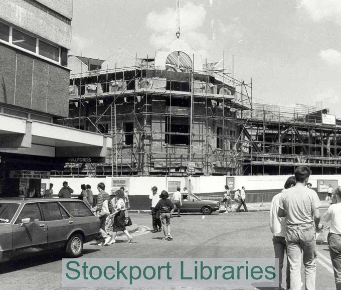 Building under construction with scaffolding, a crane, a car, and people on a street.