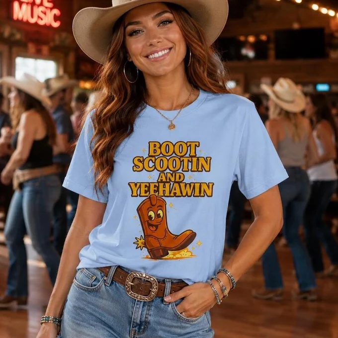 Cowgirl wearing a blue Boot Scootin And Yeehawin graphic tee with denim shorts and cowboy hat inside a lively western dance hall.