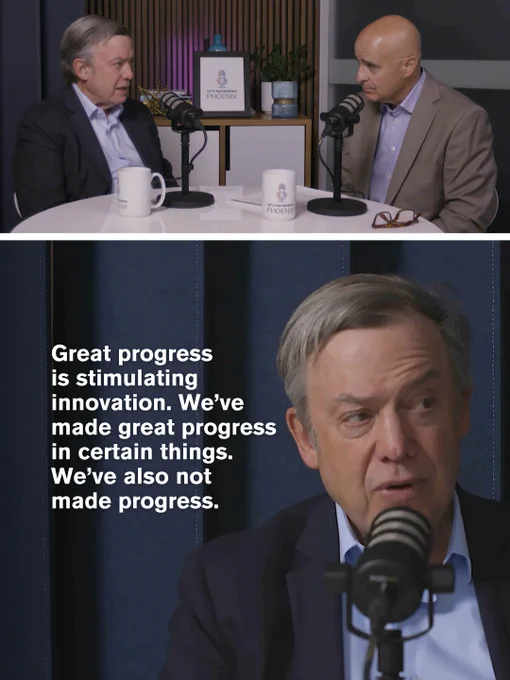 Two men in suits having a podcast discussion at a round table with microphones and  mugs in a modern studio setting.