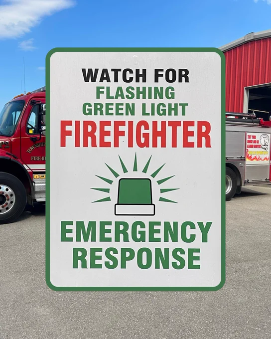Emergency response sign instructing to watch for a flashing green light, indicating a volunteer firefighter is responding to a call in a personal vehicle. In the background of the image there is a fire truck.