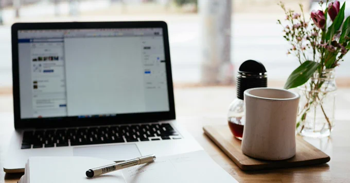 A tidy workspace featuring an open laptop showing a Facebook page, accompanied by a notepad, a pen, a coffee mug, a small glass of tea, and a vase with fresh flowers on a wooden table.