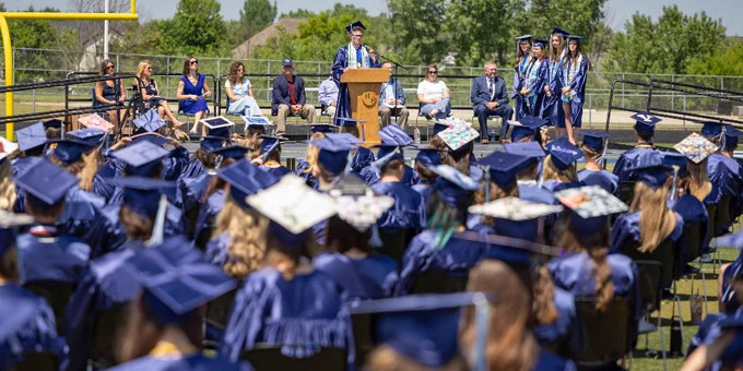 Graduate in cap and gown delivering speech at outdoor ceremony with seated audience and fellow graduates on stage.