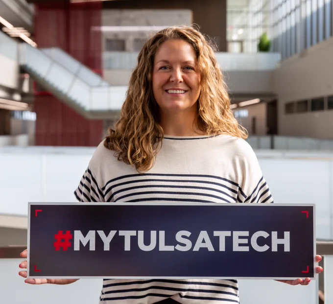 Person with curly hair holding a #MYTULSATECH sign inside a modern building with large windows and stairs.