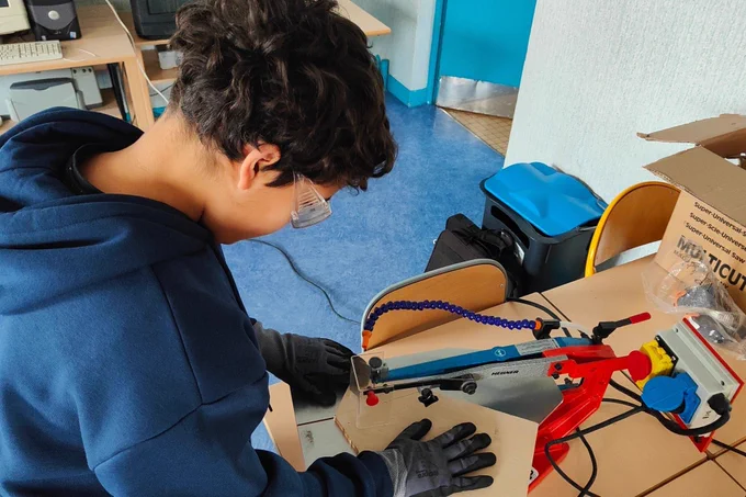 Élève équipé de lunettes et de gants de protection utilisant une scie à chantourner pour découper une plaque de bois dans un espace de fabrication en classe.