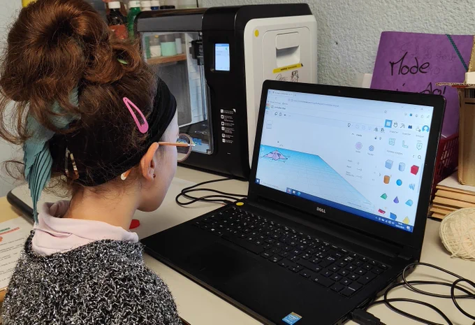 Élève utilisant un ordinateur pour modéliser un objet en 3D dans un logiciel de conception, assise devant une imprimante 3D dans une salle de classe. 