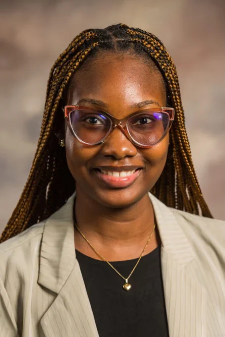 A professional headshot of Promise Oti-Okardie. She is wearing glasses, a black shirt, and a tan blazer.