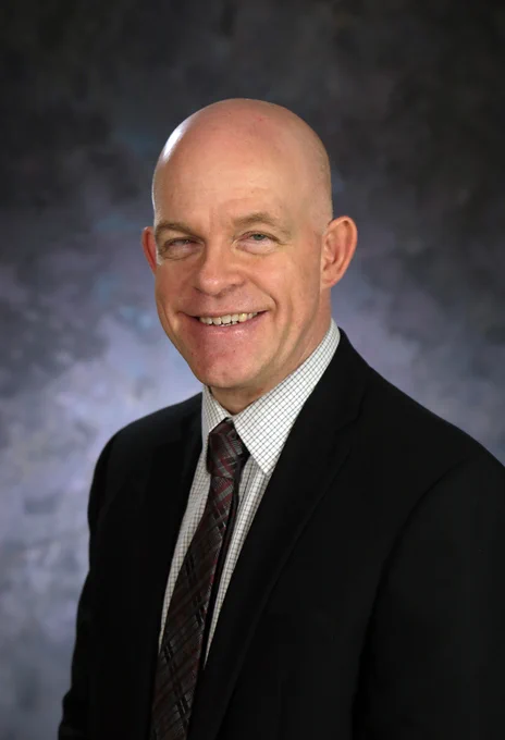 Professional portrait of James Dante wearing a dark suit, white checkered shirt, and patterned tie against a neutral gray backdrop.