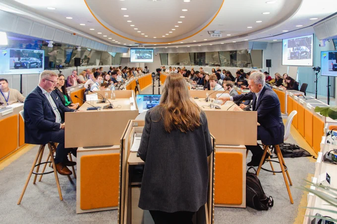 A conference room setting at the European Sustainable Energy Week with a speakers seated facing each other at the front and an audience seated in rows. There are multiple screens displaying images and presentations. The room has modern decor, featuring orange accents and a circular layout.