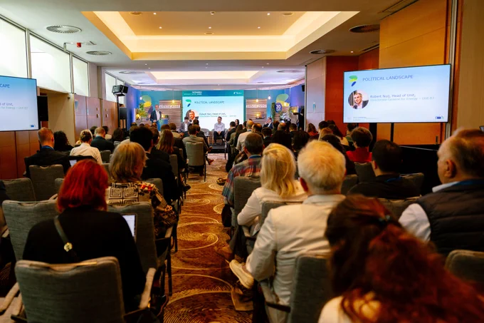 A conference room at the European Sustainable Energy Week with an audience seated and facing a presentation stage. Two screens display information, and a panel discussion is taking place at the front.