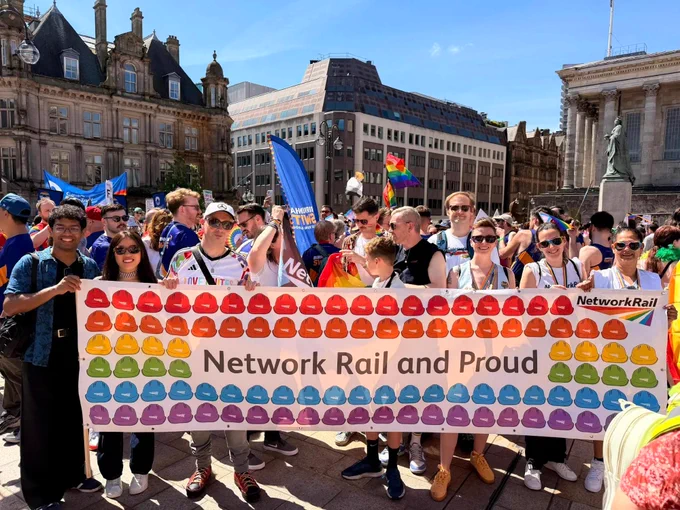 A group of people standing together in a public square, holding a large banner that reads "Network Rail and Proud" decorated with colourful hard hat icons. 