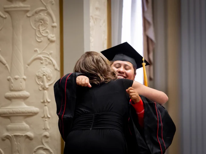 A returning adult student wearing a graduation cap and gown hugs a staff member of the Division of Continuing Studies after an emotional moment.