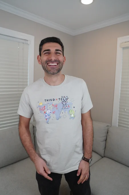Alt text: A smiling person stands indoors in front of a light gray sectional couch, wearing a light gray This Dot Labs t-shirt featuring a world map illustration with cartoon characters and location markers. They have short dark hair, a trimmed beard, dark jeans, and a wristwatch. The room has beige walls, white trim, two windows with closed white blinds, and recessed ceiling lighting, creating a bright and casual home-office setting.