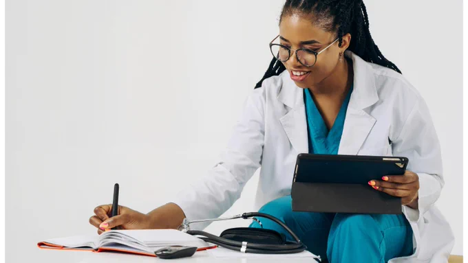 A medical professional in a white coat and teal scrubs is seated while writing in a notebook. She is holding a tablet in her other hand and wearing glasses. A stethoscope and a blood pressure cuff are placed on the table in front of her. Her expression appears focused and engaged.