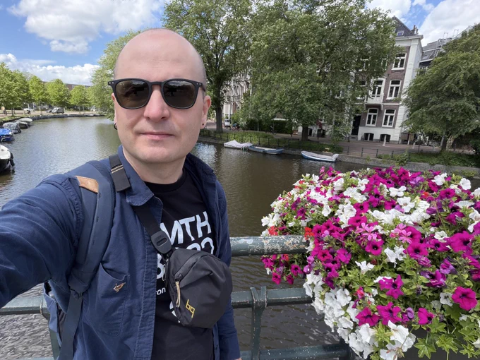 A selfie on a canal bridge in Amsterdam, with pink and white flowers in a railing flower box in the foreground, moored boats and classic Dutch brick buildings visible in the background.