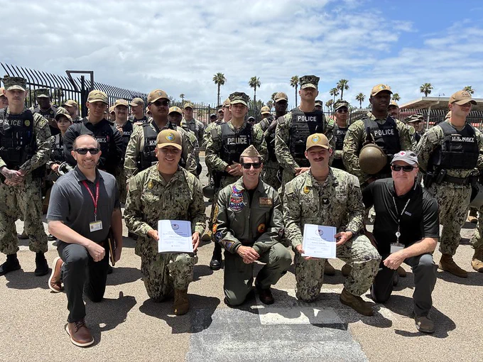 A group of people in military uniforms and personnel in military police vests are posing outdoors. Two individuals at the front hold certificates. The scene includes a clear sky and palm trees in the background.
