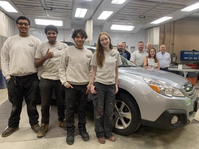 A group of young adults, staff and high school students around a silver Subaru outback in an automotive service garage  happy about the car renovation. 