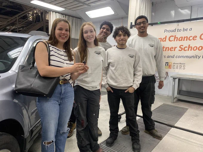 The lady who gave their car for car renovation holding a key happy about the final look of the car, while the students who helped renovate it is also in the picture 