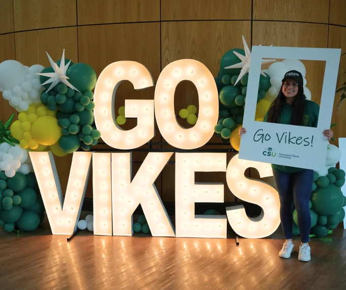 CSU student taking a photo in front of illuminated Go Vikes sign and holding a polaroid cutout frame. 