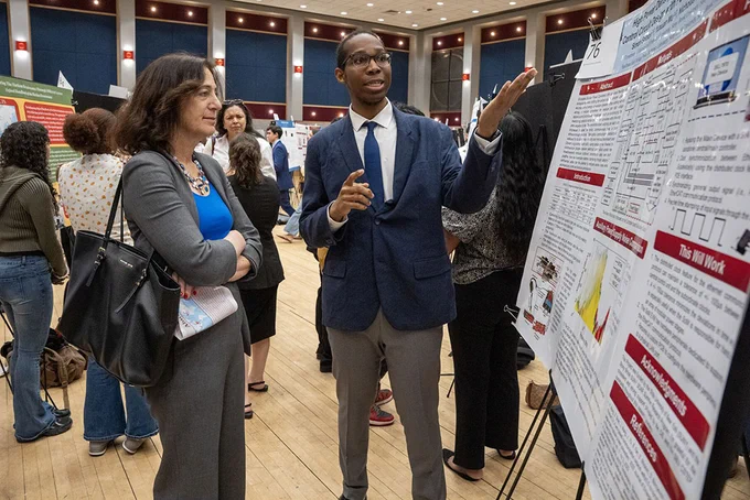 A person in a suit explains research displayed on a poster board to another individual in a conference hall. Several attendees and poster presentations are visible in the background.