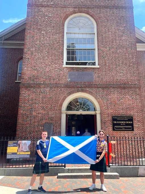 Young men wearing kilts and holding the Scottish flag in front of the Old North Church. 