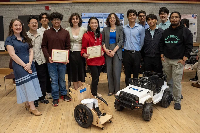 A group of people is standing indoors on a wooden floor. Two individuals are holding certificates. In front of the group, there are two robotic vehicles, one resembling a small car and the other a wooden cart. Posters are displayed on the wall in the background.