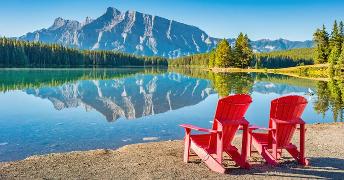 Two red Adirondack chairs overlook Two Jack Lake in Banff National Park on a sunny morning.