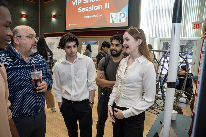 A group of people stand in a room with display boards and a large model rocket. They appear to be engaged in discussion. One person is drinking a beverage. A sign in the background reads "VIP Showcase Session II.